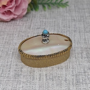 May include: An oval-shaped, antique jewelry box with a gold-toned exterior and a pearlescent lid. The lid is adorned with a small, ornate silver-colored knob topped with a turquoise stone. The box has a textured, ridged border.