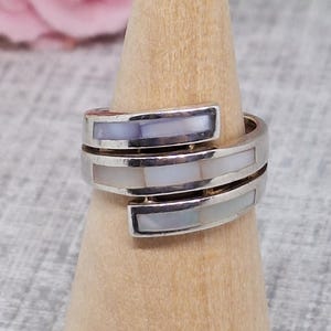 May include: A silver-toned wrap ring with rectangular mother-of-pearl inlays. The ring is displayed on a wooden ring holder. The ring's design features a spiral shape with three sections of inlaid material.