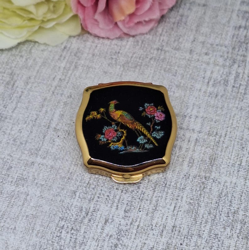 May include: A vintage compact with a gold-tone frame and a black enamel top. The top features a colorful illustration of a pheasant perched on a flowering branch. The compact has a rectangular shape with curved edges.