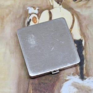 Op de afbeelding: Een vierkant zilveren compact met een klein gegraveerd logo met de tekst "Made in England" en "COTY".