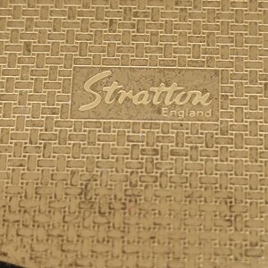 May include: A gold-toned compact with a woven pattern. The compact features the name "Stratton England" in a rectangular frame. The compact has a curved edge and a shiny finish.