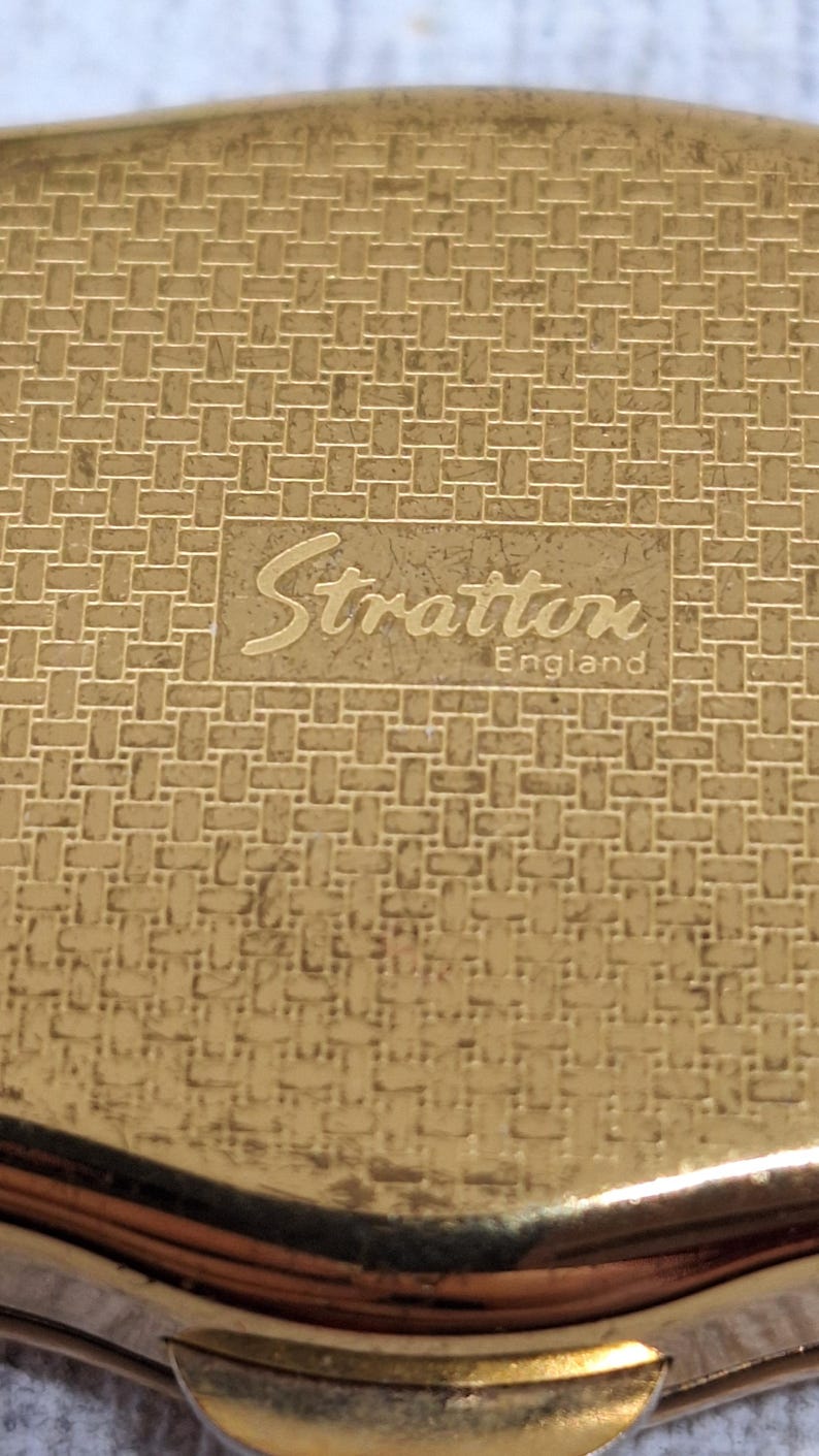 May include: A gold-toned compact with a woven pattern. The compact features the name "Stratton England" in a rectangular frame. The compact has a curved edge and a shiny finish.