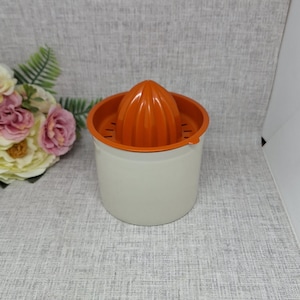 Op de afbeelding: Een vintage citruspers met een feloranje ruimer en rand, geplaatst in een crèmekleurige container. De pers is ontworpen voor het gemakkelijk persen van citrusvruchten. De afbeelding toont de pers op een neutrale achtergrond.
