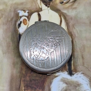 May include: Silver compact with an engraved design of a woman in a dress with flowers. The compact has a striped pattern and a blank rectangular space for engraving.