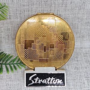 Puede incluir: Un compacto redondo dorado con un patrón geométrico y la palabra "Stratton" en la parte inferior.