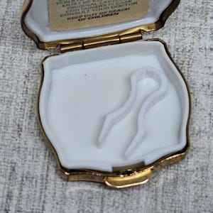 May include: Open, gold-toned pill box with a white interior and two white plastic applicators. The lid has a small printed label with text. The box is open, revealing the contents.