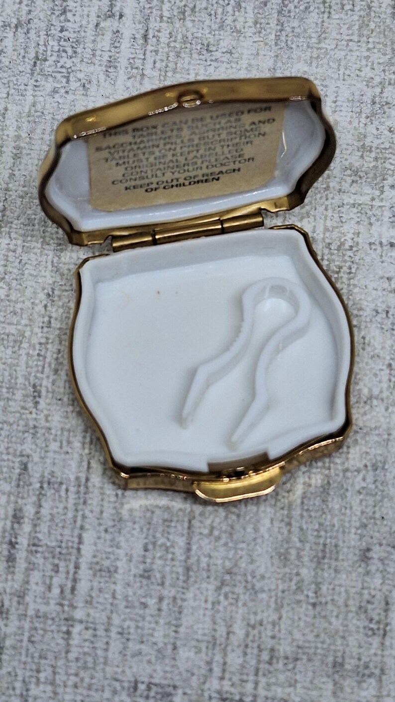 May include: Open, gold-toned pill box with a white interior and two white plastic applicators. The lid has a small printed label with text. The box is open, revealing the contents.