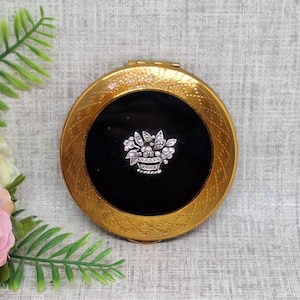 Peut inclure: Un compact doré vintage avec un centre noir. Le compact présente un motif floral décoratif en strass. Le compact est rond et possède une bordure texturée.
