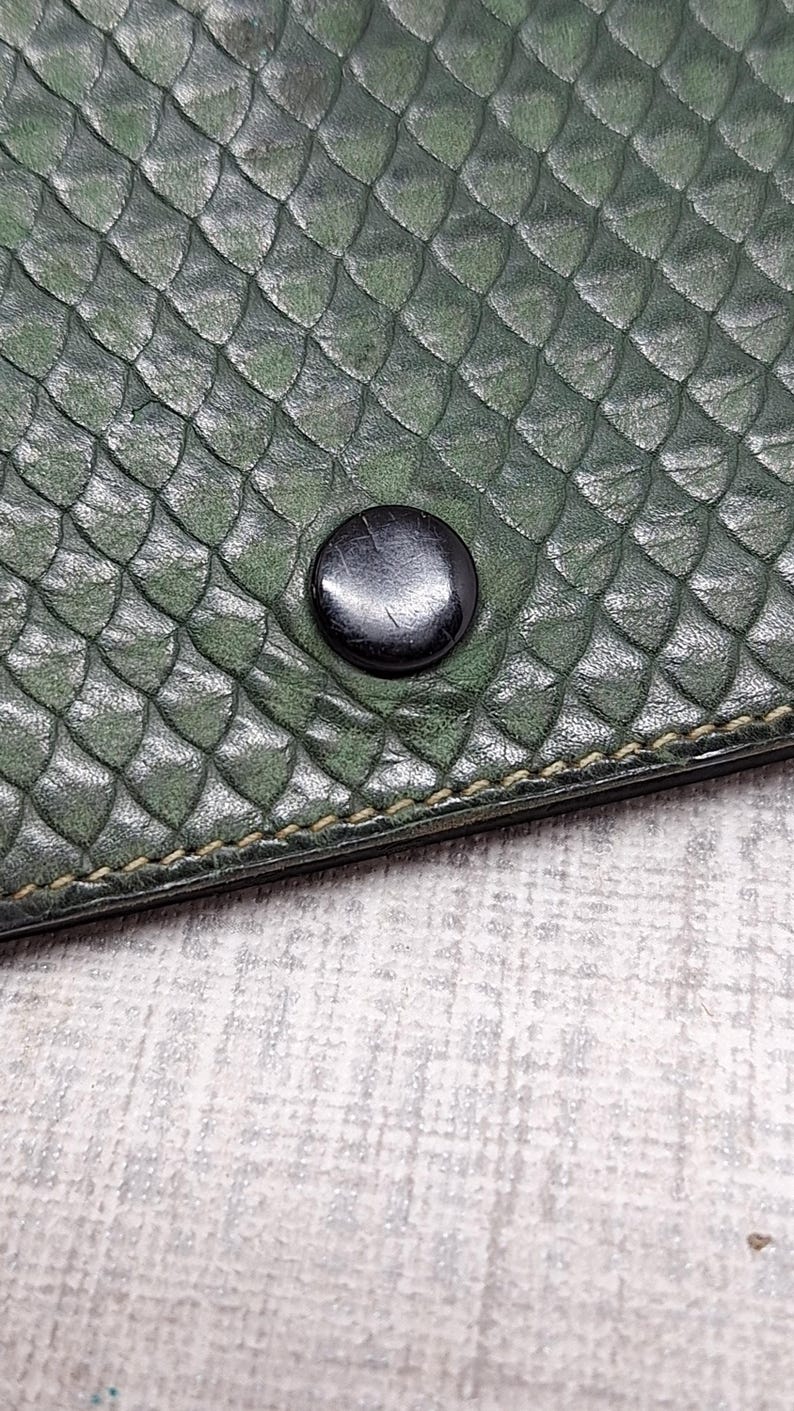 Pu&ograve; includere: Primo piano di un articolo in pelle verde scuro con una trama a scaglie. Un bottone nero &egrave; centrato sull'articolo. La cucitura &egrave; visibile lungo il bordo dell'articolo.