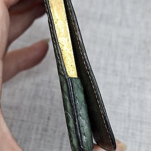 Pu&ograve; includere: Un portafoglio vintage aperto, che rivela un interno color oro. L'esterno &egrave; in pelle verde scuro con un motivo strutturato. Il portafoglio &egrave; tenuto aperto, mostrando l'interno e le cuciture dettagliate.
