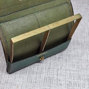 Pu&ograve; includere: Portafoglio in pelle verde aperto con cornice in ottone. Il portafoglio ha un esterno strutturato e una chiusura a scatto. La scritta dorata all'interno recita "for 1932 from b."