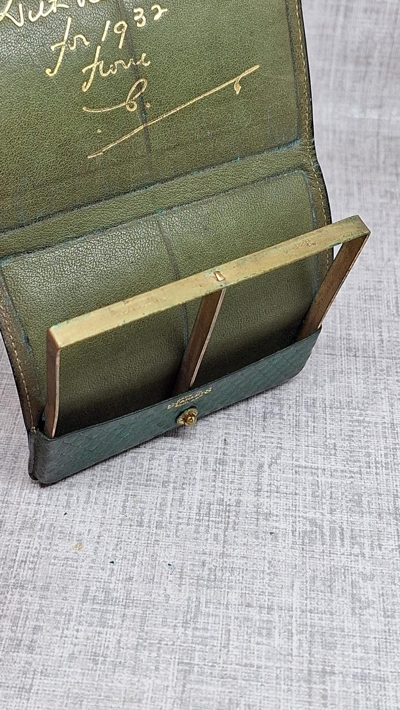 Pu&ograve; includere: Portafoglio in pelle verde aperto con cornice in ottone. Il portafoglio ha un esterno strutturato e una chiusura a scatto. La scritta dorata all'interno recita "for 1932 from b."