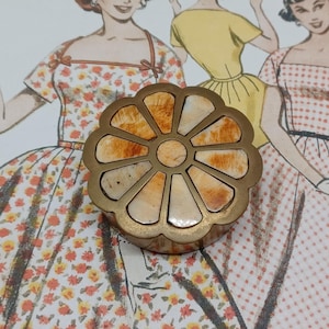 May include: A brass trinket box with a floral design. The box is made of brass and has a circular shape. The top of the box is decorated with a flower design made of inlaid stone. The flower has a light brown centre and petals in shades of orange and brown.