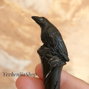 Pode incluir: Uma estatueta de corvo preto empoleirada em cima de uma base esculpida de cor escura. O corvo tem texturas de penas detalhadas e um olhar focado. A base tem um design em espiral. O texto "YevheniiaShop" é visível.