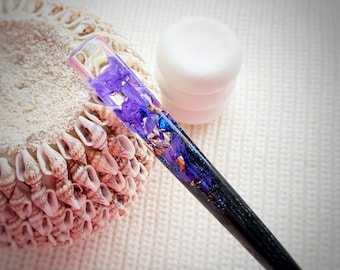 Puede incluir: Un palillo decorativo para el cabello con un mango negro y una parte superior de resina transparente llena de flores moradas y copos dorados. El accesorio para el cabello se muestra junto a una concha y una pila de objetos blancos.