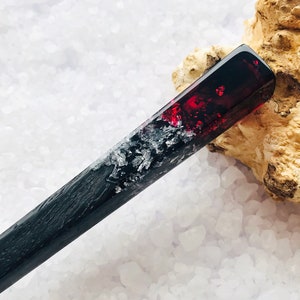 May include: A black and red hair stick with a red resin section containing silver glitter. The stick is resting on a white surface.