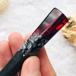 May include: A black and silver resin hair stick with a red and silver resin top. The stick is about 6 inches long.