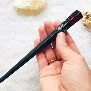 May include: Black and red resin hair stick with a pointed end. The stick is held in a hand.
