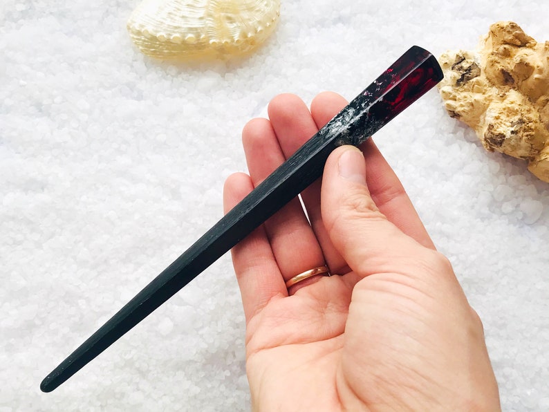 May include: Black and red resin hair stick with a pointed end. The stick is held in a hand.