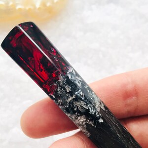 May include: A black and red resin hair stick with silver flecks. The stick is held in a hand.