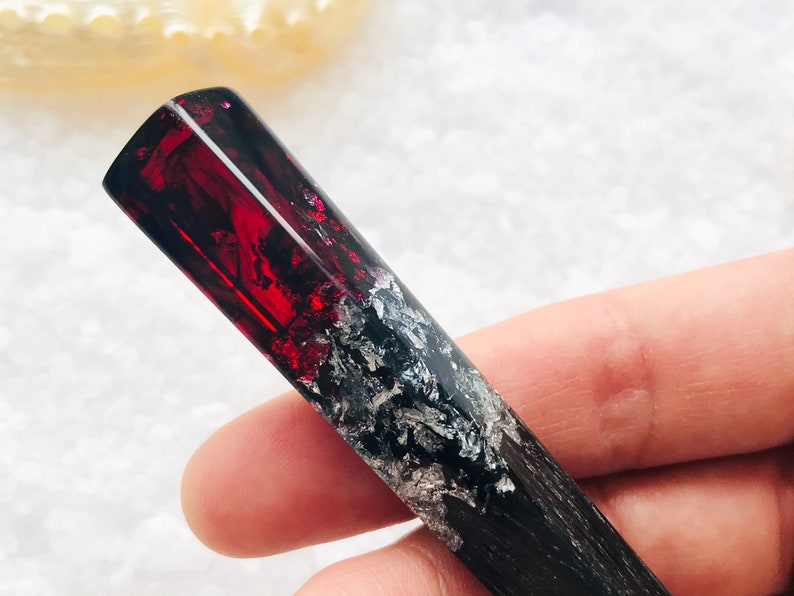 May include: A black and red resin hair stick with silver flecks. The stick is held in a hand.