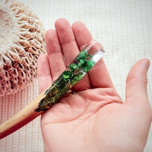 Pode incluir: Um pau de cabelo de madeira com uma seção de resina verde e dourada contendo flores e folhas prensadas.