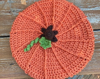 Puede incluir: Una calabaza de ganchillo naranja hecha a mano con un tallo marrón y una hoja verde. La calabaza es redonda con líneas verticales que irradian desde el centro. La calabaza está sobre una superficie de madera.