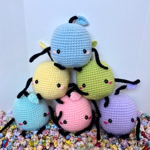 May include: A group of six crocheted plush toys in various colours, including light blue, yellow, green, pink, and purple. Each toy has a round body, black eyes, and a small mouth. The toys are arranged in a pyramid shape, with the largest toy at the top and the smallest toy at the bottom.
