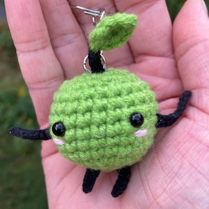May include: A green crocheted plushie with black arms and legs and a small green leaf on top. The plushie has a cute face with black eyes and pink cheeks. It is attached to a silver keyring.
