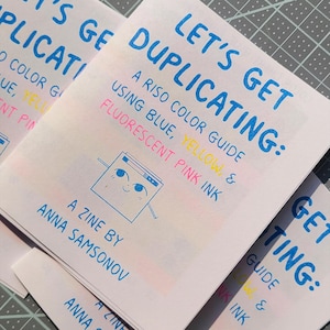 Könnte beinhalten: Ein Zine mit dem Titel Let's Get Duplicating: A Riso Color Guide Using Blue, Yellow, & Fluorescent Pink Ink von Anna Samsonov. Das Cover zeigt blauen Text, eine kleine illustrierte Figur sowie gelbe und rosa Akzente auf weißem Papier.