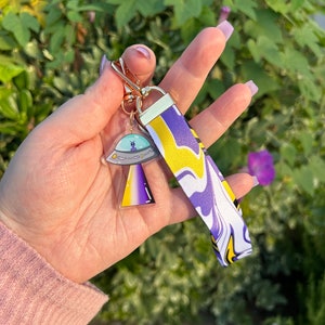 May include: A keychain with a gold-colored clasp and a clear acrylic charm featuring a cartoon alien in a spaceship. The keychain also includes a wrist strap with a purple, yellow, and white swirl pattern.