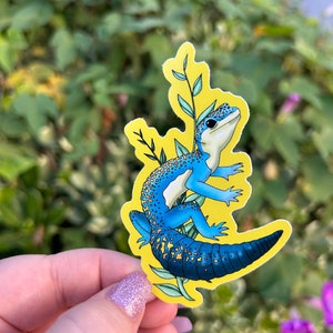 May include: A sticker featuring a blue and white gecko with a yellow background and green leaf accents. The gecko has a long tail and is surrounded by green leaves. The sticker is held by a hand with glittery nail polish.