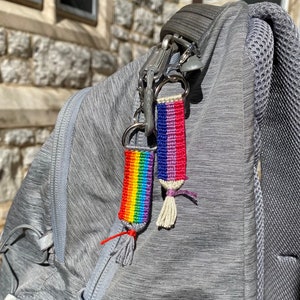May include: A gray backpack with two rainbow and purple woven keychain tassels attached to a silver key ring.
