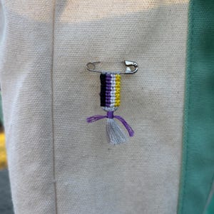 May include: A silver safety pin holds a small, handmade charm. The charm features a striped design in black, purple, white, and yellow, with a gray tassel and purple accents. The charm is attached to a cream-colored canvas.