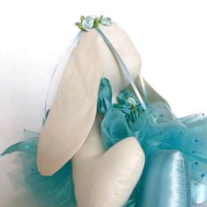May include: A white stuffed bunny wearing a blue tulle tutu with white and green floral accents. The bunny is wearing blue satin ballet shoes.