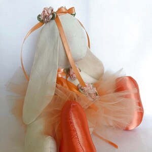 May include: A white fabric bunny wearing a peach tulle tutu and orange satin shoes. The bunny has a peach satin ribbon headband with pink fabric flowers.