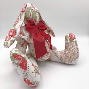 May include: A patchwork fabric bunny with a red bow. The bunny is sitting on a white surface. The fabric is a mix of white, green, and red with a floral pattern.