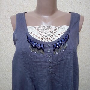 May include: A gray sleeveless top with a white crocheted neckline and a beaded embellishment. The top has a button closure down the front.