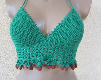 May include: A green crochet halter top featuring a triangular bodice with a scalloped edge and a midriff section with an openwork pattern. The bottom hem is decorated with a series of small, dangling brown crocheted strawberry-shaped charms.