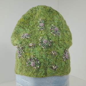 May include: A green, textured hat decorated with floral details. The hat features clusters of small, pale purple roses, white pearls, and green beads. The hat has a rounded shape and a soft, fuzzy texture.