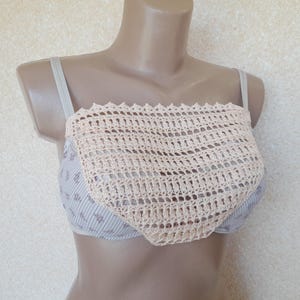 May include: A beige crochet top with a square design is displayed on a mannequin. The crochet top is worn over a light blue and white striped bra with a floral pattern. The bra has thin straps.