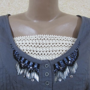 May include: A gray sleeveless top with a white crocheted trim and a beaded embellishment. The embellishment features silver and blue beads.