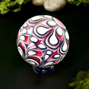 May include: A decorative glass sphere with a swirling pattern of red, white, and dark blue teardrop shapes. The sphere sits on a dark blue base, and the background is black with green moss and small stones.