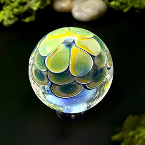 May include: A clear glass paperweight with a floral design. The paperweight features a spherical shape with green, yellow, and blue petal-like patterns. The base is dark blue. The background is black with green moss and small stones.