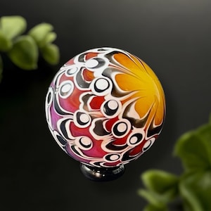 May include: A colorful glass orb with a black base. The orb features a vibrant design with red, yellow, white, and black patterns. The glass art piece is round and appears to be handmade.