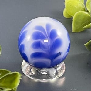 May include: A blue and white glass marble with a floral pattern. The marble is displayed on a clear glass stand. The design features swirling blue patterns against a white background. The marble is a sphere.