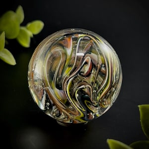 May include: A clear glass marble with swirling patterns of brown, yellow, white, and black. The marble has small air bubbles and a rounded shape. The background is black, with green leaves in the corners.