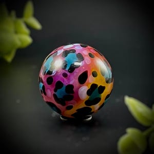 May include: A colorful glass sphere with a vibrant pattern of black spots on a background of pink, blue, orange, and yellow. The sphere is resting on a small metal stand, with a dark background and green foliage in the corners.