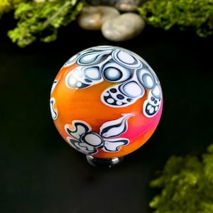 May include: A decorative glass orb with a vibrant orange and pink gradient. The surface features white and black floral and abstract patterns. The orb is set on a black base, showcasing a unique, artistic design.