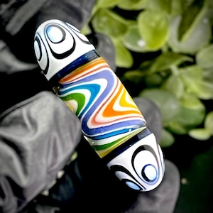 May include: A colorful glass bead with a swirling pattern of blue, green, orange, and white. The bead has black and white circular designs on each end. The bead is held in a black gloved hand.